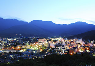 夜が近いがまだ青白い空を背景に、手前の暗い山々に囲まれた別府市街地。建物から漏れるオレンジや薄緑の光の中に無数の湯けむりが上がり、幻想的な風景の写真